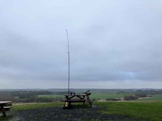 The top of a hill with a view over typical British countryside, complete with a typical British grey sky. In the foreground is a picnic bench, with a bike propped up on one side and a very tall pole rising up on the other. Just about visible on the table is my radio.