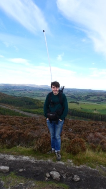 A photograph of me, with a valley and hills in the background. I am wearing a rucksack with a large white vertical antenna attached to the back of it.