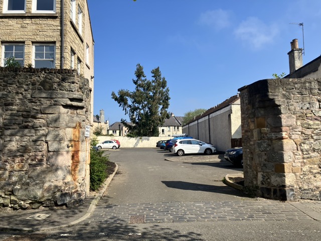 Very thick medieval-looking stone walls with a gap for a gate in them. Through the gap is a car park and a modern housing development.